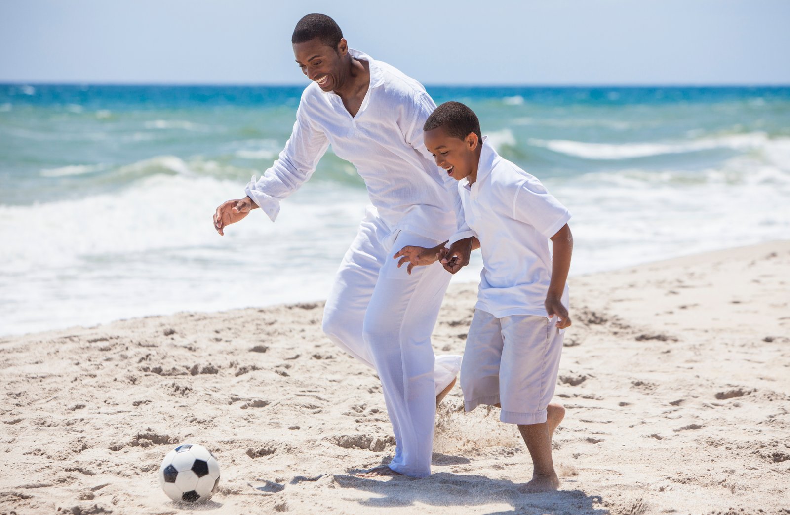 African American family of father and son, man &amp; boy child, having fun playing football soccer in the sand on a sunny beach